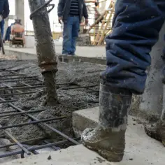 Betoniarka wylewa beton na zbrojenie, pracownicy w gumowcach nadzorują proces.