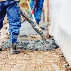 Pracownicy w niebieskich kombinezonach i gumowcach wylewają beton na zbrojenie.