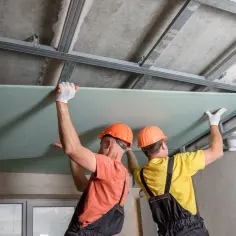 Dwóch budowlańców w kaskach montuje płytę gipsowo-kartonową do metalowego stelaża sufitu.