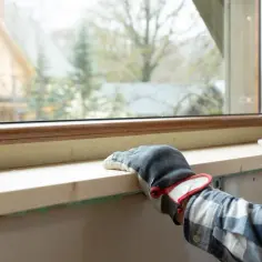 Ręce w rękawiczkach montują drewniany parapet wewnętrzny. Zastanawiasz się, ile za montaż parapetów wewnętrznych?