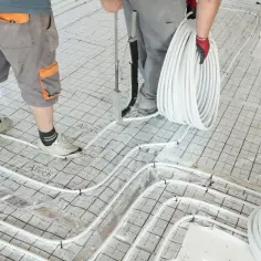 Instalacja ogrzewania podłogowego: pracownicy układają białe rury na siatce.