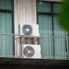 Dwa białe klimatyzatory zamontowane na ścianie budynku, na tle okien z zielonymi zasłonami.