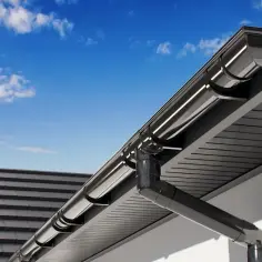Szary system rynnowy zamontowany na dachu domu pod błękitnym niebem z chmurami.