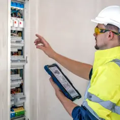 Elektryk w kamizelce odblaskowej i kasku sprawdza schemat na tablecie, analizując, ile kosztuje uzbrojenie skrzynki elektrycznej.