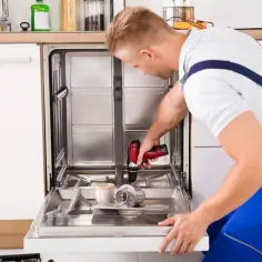 Mężczyzna w niebieskim kombinezonie używa wkrętarki do naprawy otwartej zmywarki.