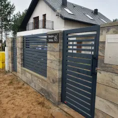 Nowoczesne ogrodzenie z ciemnoszarych paneli i słupków z kamienia, z widocznym numerem posesji.