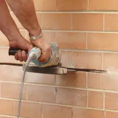 Ręce trzymają szlifierkę kątową, która przecina ceglaną ścianę.